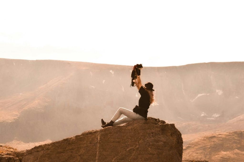 A woman sits on a rock in an arid landscape, holding a cat aloft under the sunlight.