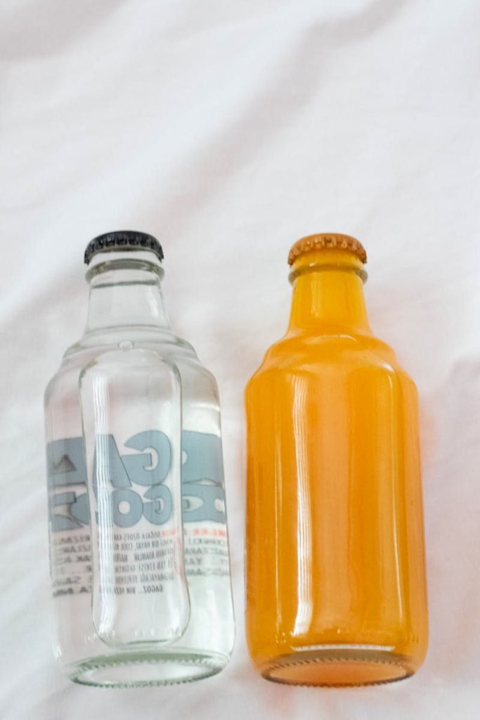 Two glass soda bottles, clear and orange, placed on a white surface.