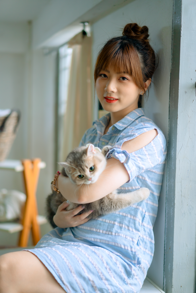 An Asian woman in a blue dress holding a cat indoors with natural light.