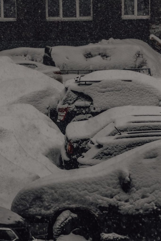 Heavy snow blankets parked cars in a quiet urban setting on a winter day.