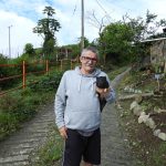 A senior man in a hoodie holding a piglet while standing on a rural path with greenery around.