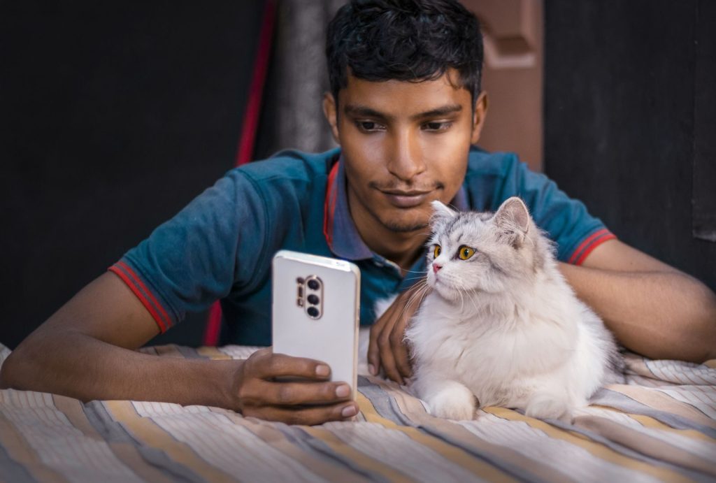 A young man lying with his Persian cat while using a smartphone indoors.