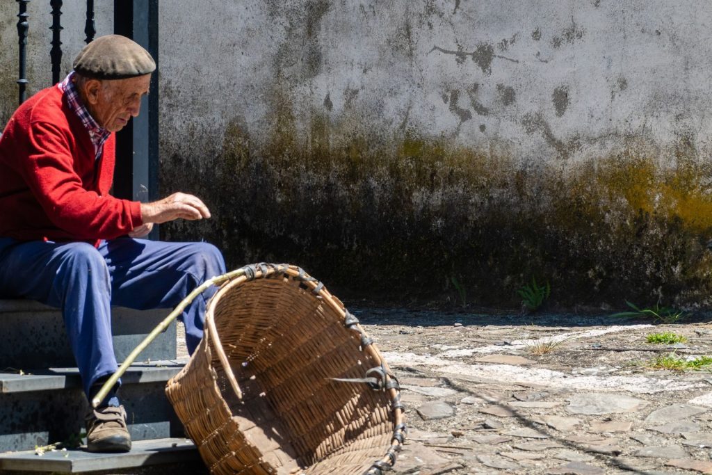 Elderly man in red sweater sitting outdoors with a large basket on city steps.