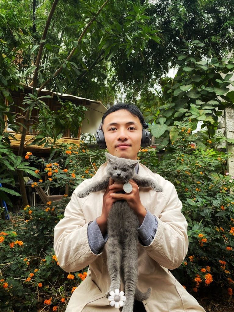 A young man in a coat holds a cat, surrounded by lush greenery and flowers.
