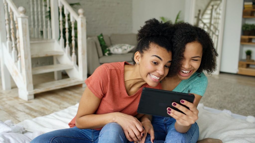 Two young women laughing and enjoying on a tablet, indoors, relaxed atmosphere.