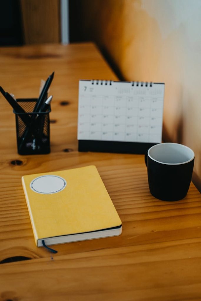 Warm wooden desk with calendar, yellow notebook, and black mug creating a cozy workspace vibe.