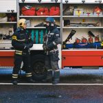 Two firefighters in full gear stand by a fire truck discussing strategy. Open equipment compartments visible.