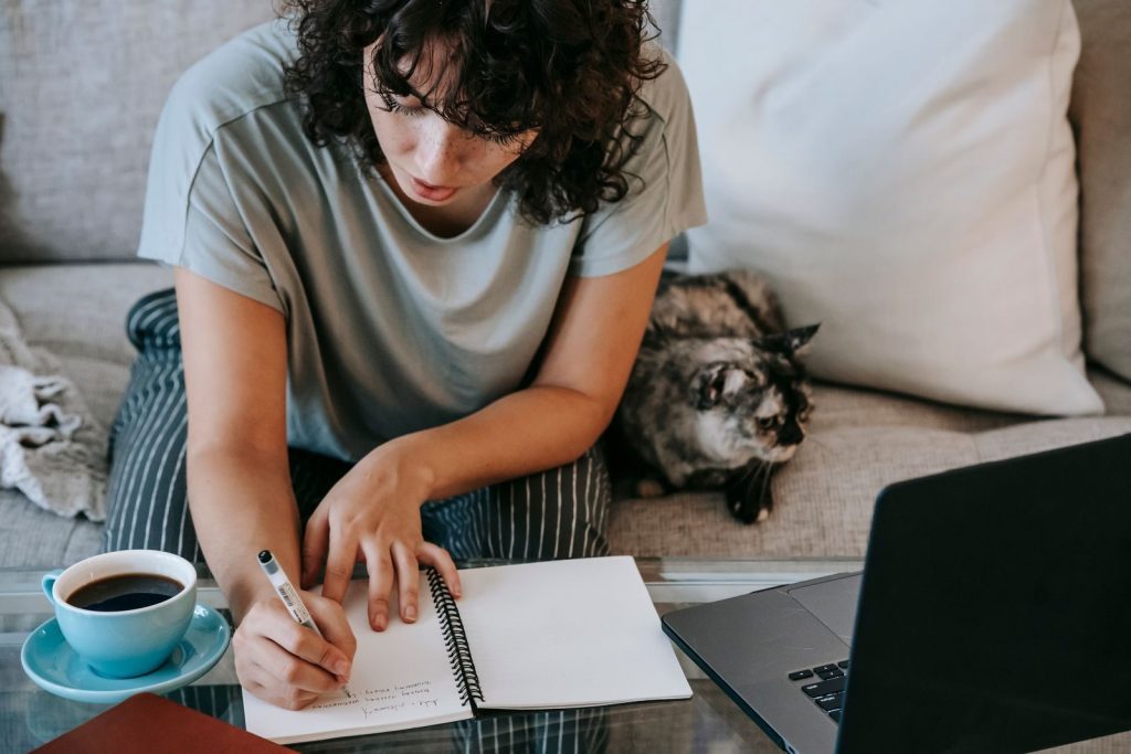 From above of crop concentrated young female student taking noted in copybook while doing homework assignment at table with laptop and cup of coffee sitting on sofa near cute cat