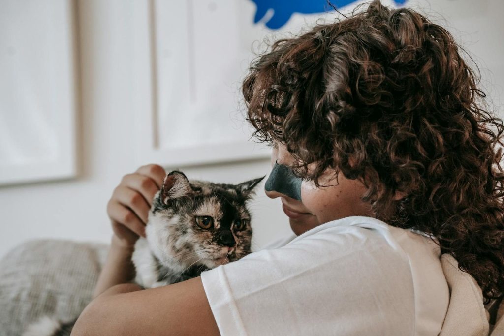 Side view of content young lady with dark curly hair and charcoal cleansing strip on nose smiling while hugging loyal cat at home