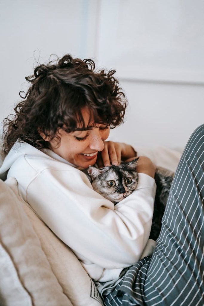 Side view of happy adult female embracing and stroking attentive cat on cozy sofa at home