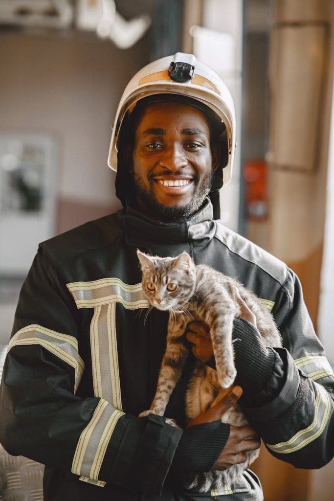 Firefighter in gear smiling while holding a rescued cat indoors.