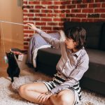 A woman sitting on the floor playing with a black cat using a toy in a cozy living room.