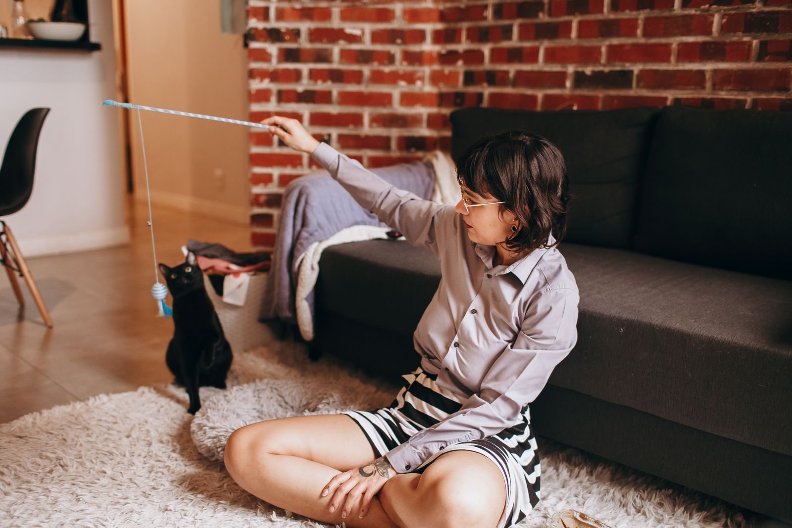 A woman sitting on the floor playing with a black cat using a toy in a cozy living room.