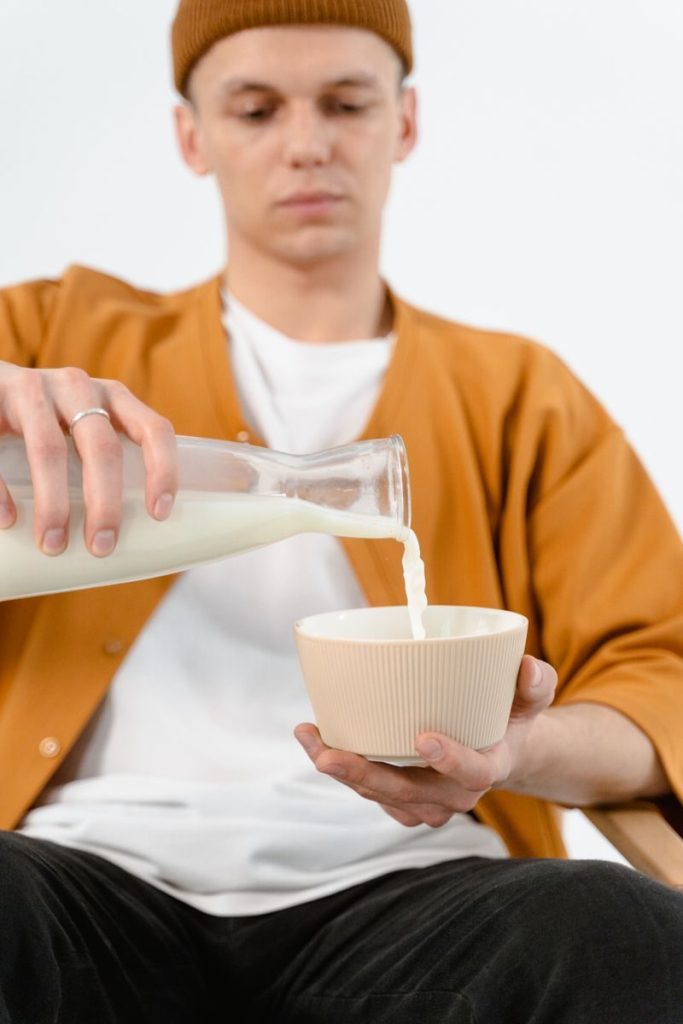 Young man in knit cap pouring milk into a ceramic bowl against a white background.