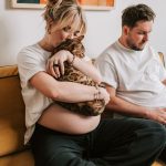 Cozy scene of a pregnant woman with a cat, while her partner works on a laptop on the sofa.
