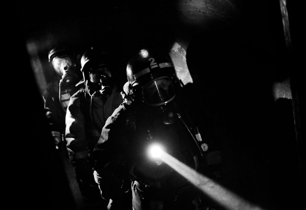 Grayscale image of firefighters navigating darkness with protective gear and a flashlight.