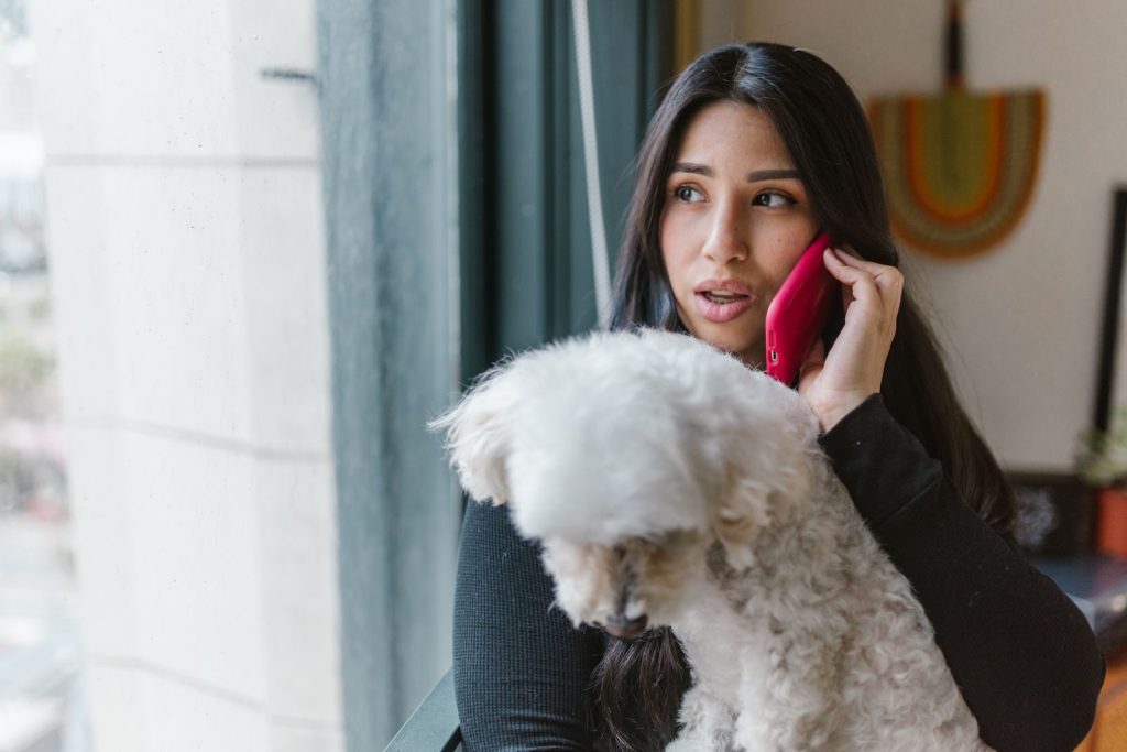 Woman holding a dog while talking on a smartphone by a window indoors.
