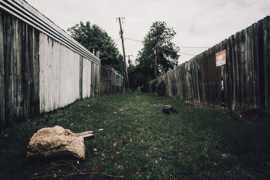 Grassy alley with wooden and metal fences on both sides, showcasing urban landscape.