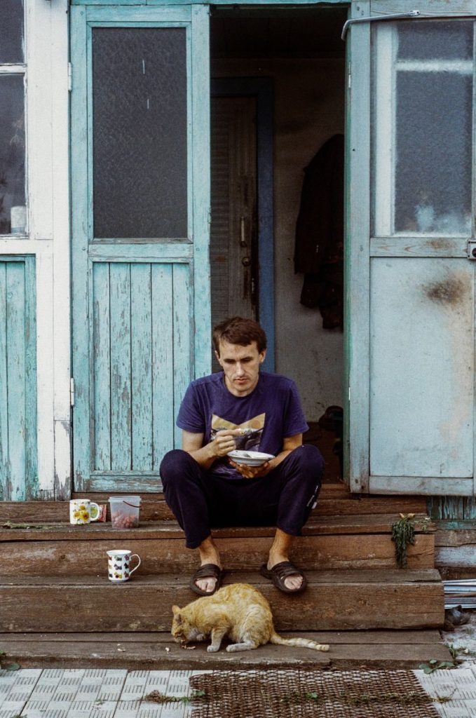 A man sits on steps eating, with a cat beside him, outside a rustic house with teal doors.