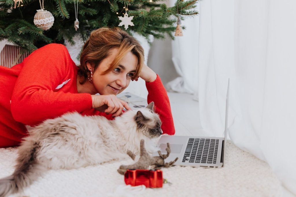 Woman in red sweater relaxing with a cat by a Christmas tree and laptop, conveying festive coziness.