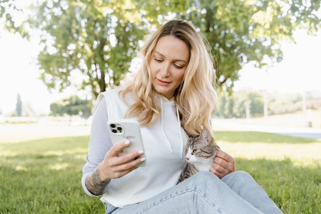 A woman using a smartphone while sitting on grass with a cat in a sunny park.