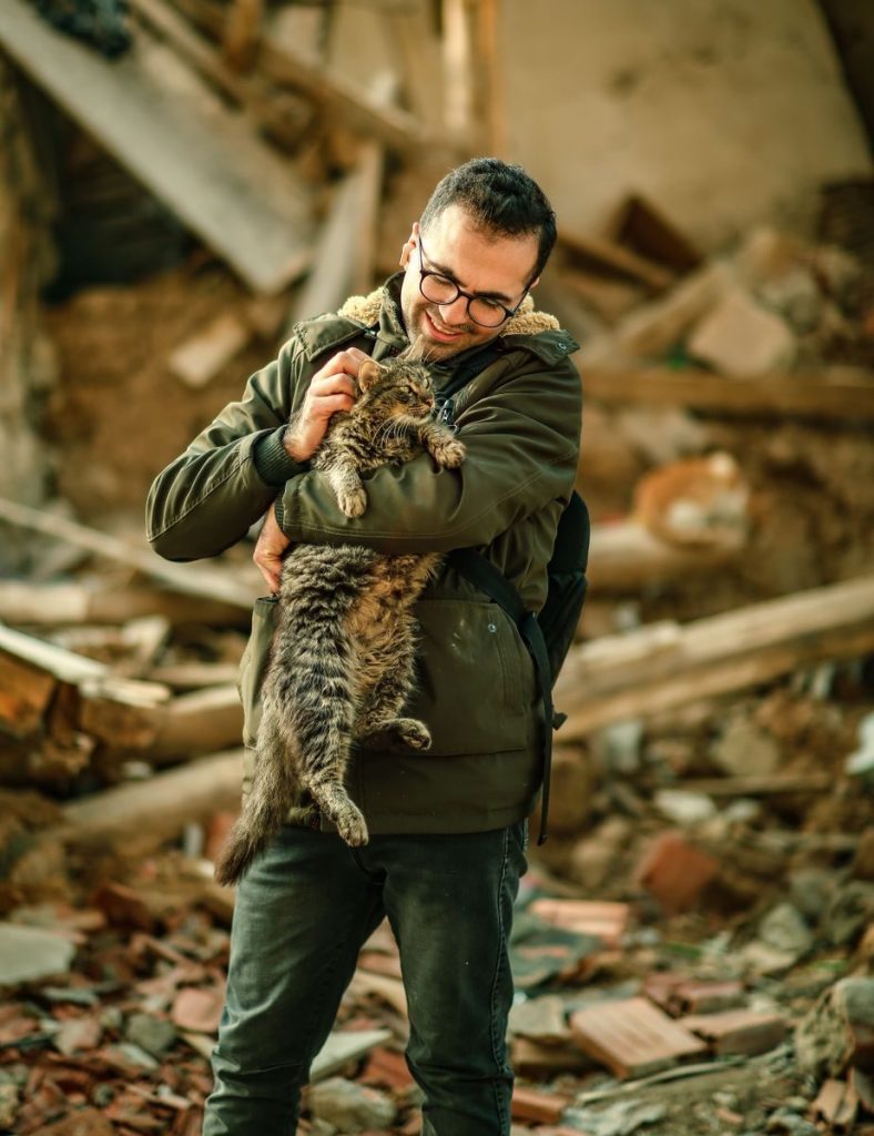 A man wearing glasses joyfully holds a tabby cat in a dilapidated urban setting.