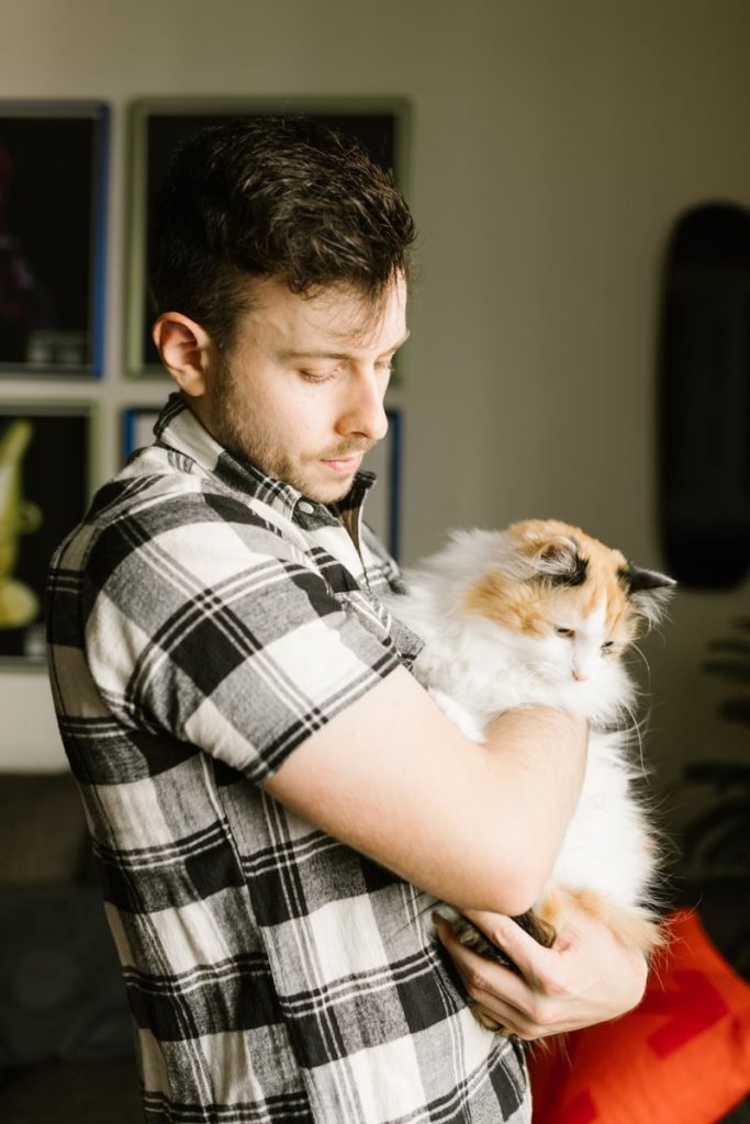 Young man in plaid shirt tenderly holding a fluffy cat indoors, expressing warmth and connection.