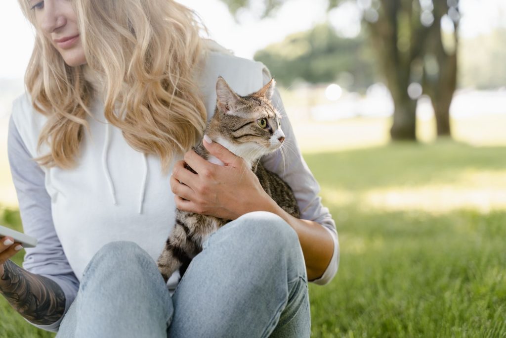 Young woman with blonde hair holding a cute tabby cat while sitting on a grassy lawn.